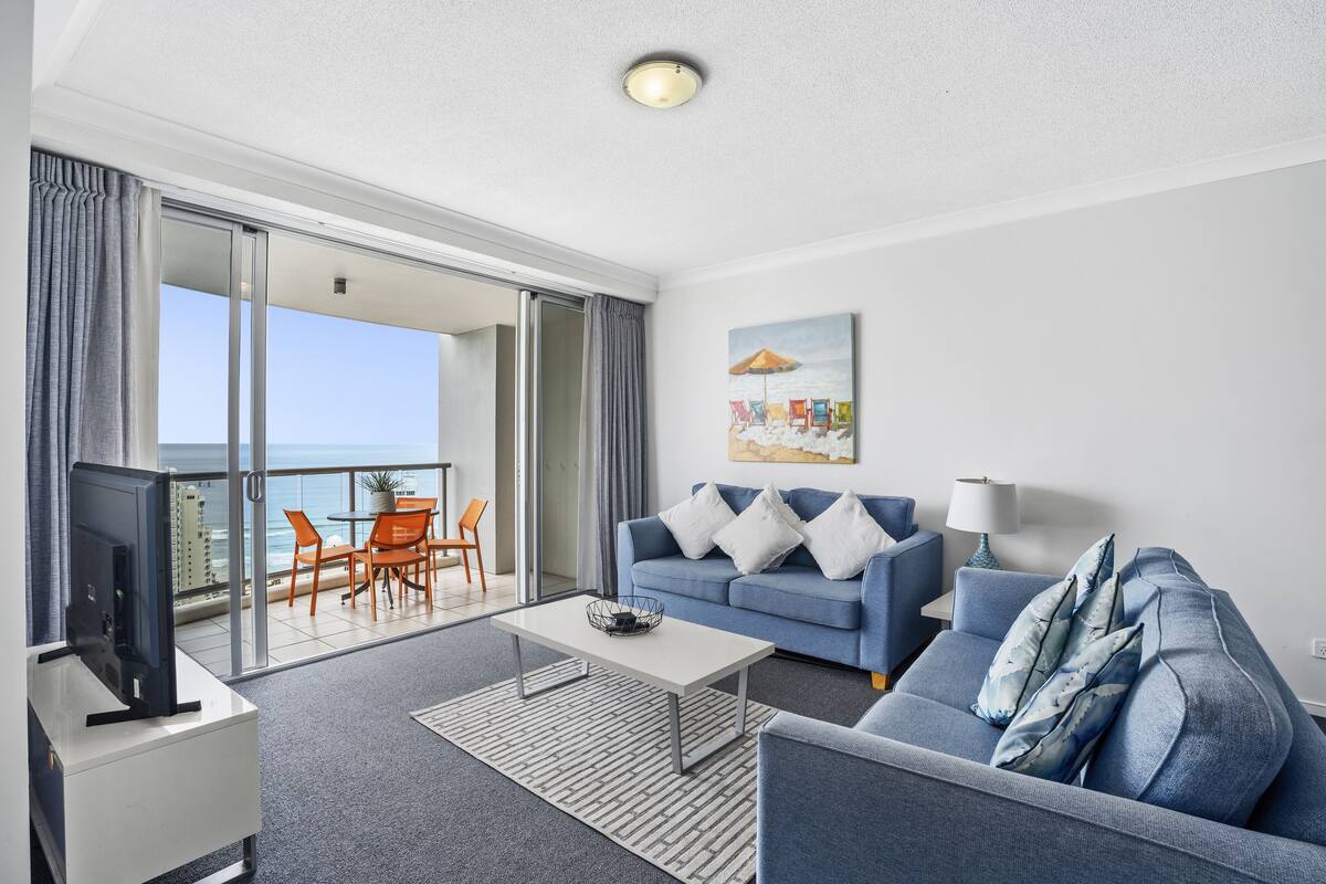 Un salon moderne avec un canapé, une table basse et un téléviseur, un balcon offrant une vue sur la mer et un tableau accroché au mur.