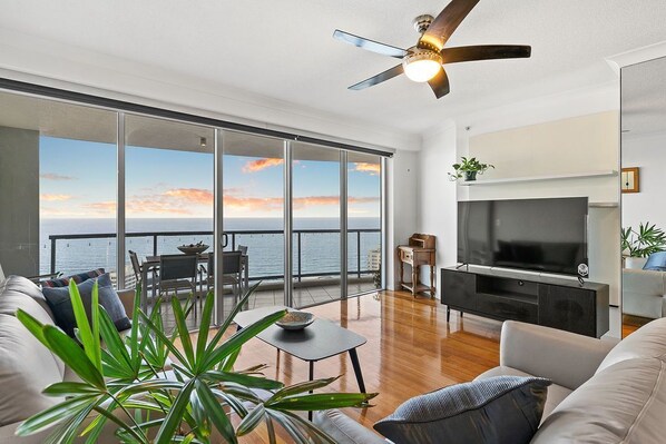 A modern living room with a large TV, a ceiling fan, and a balcony with a view of the sea.