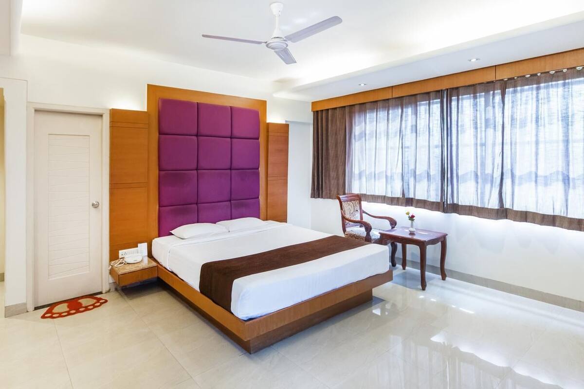 Ein modernes Hotelzimmer mit einem großen Bett, einem Holz-Kopfteil, einem Deckenventilator, einem Fenster mit Vorhängen und einem kleinen Tisch mit Stuhl.