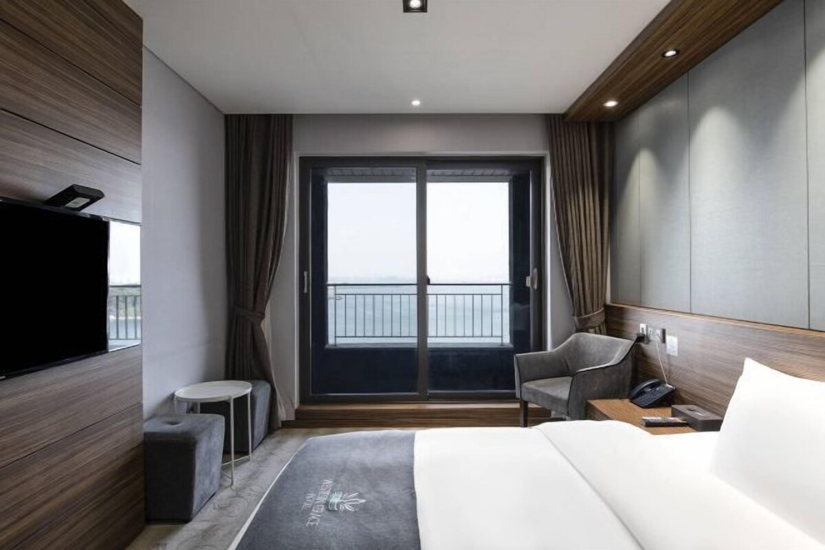 Una habitación de hotel moderna con una cama grande, un televisor en la pared izquierda, una mesita, una silla y un balcón con vistas.