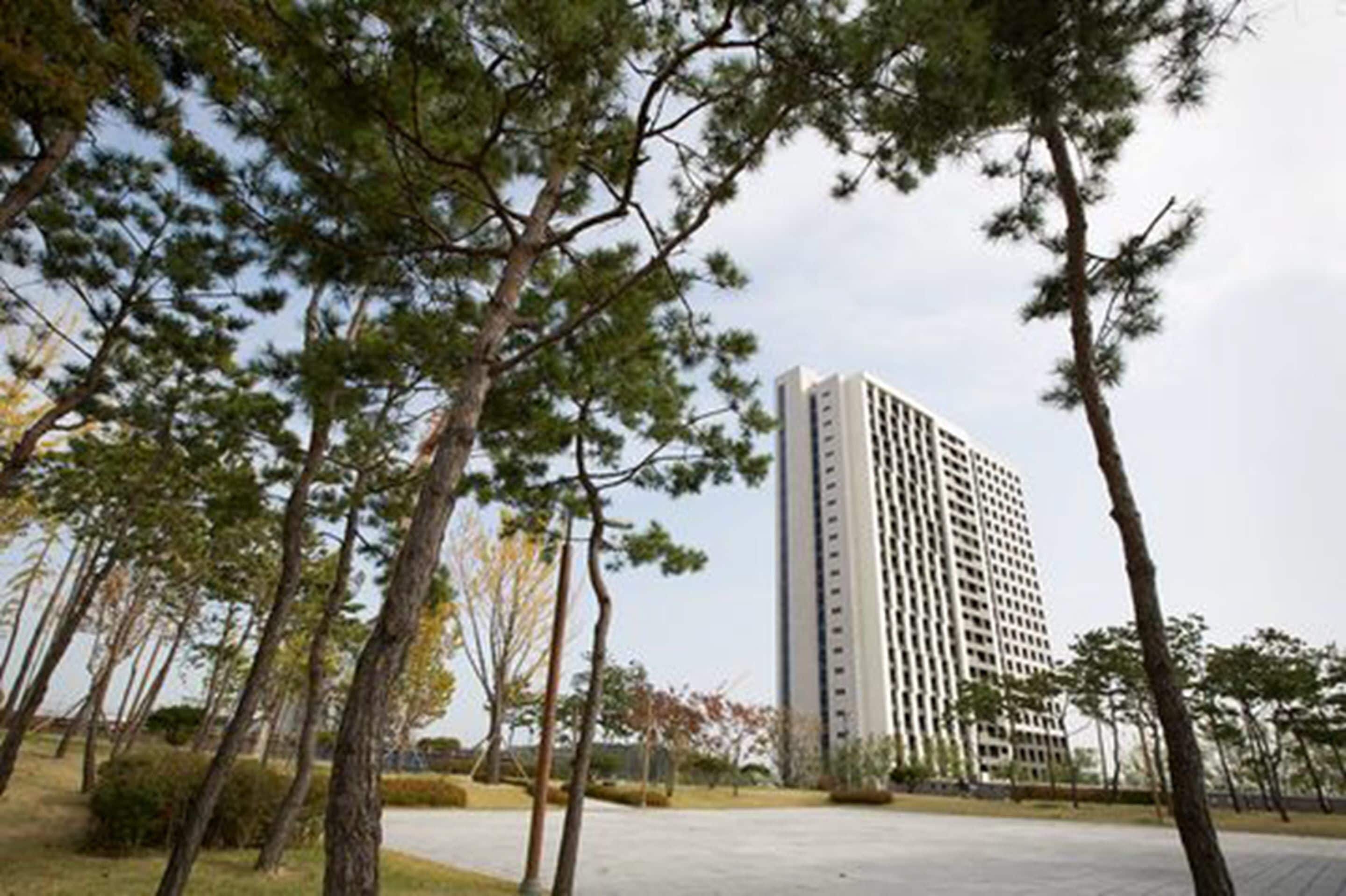 A tall, modern building with a rectangular shape and a flat roof, surrounded by a landscaped area with trees and a paved walkway.