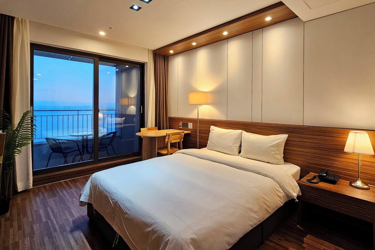 Habitación de hotel con una cama grande, un escritorio con lámpara, una silla y vistas al mar a través de una puerta corredera.