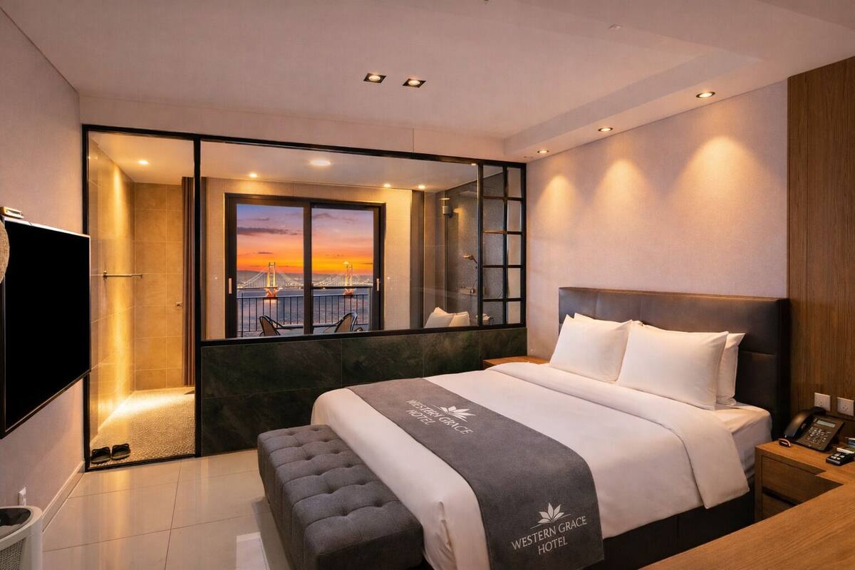 Una habitación de hotel moderna con una cama grande, vista a un atardecer sobre un puente y una mesita de noche de madera con un teléfono.