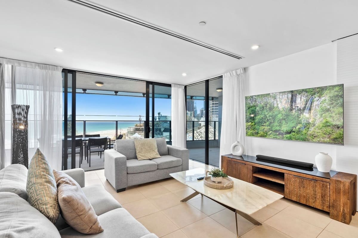 A modern living room with a grey sofa, a coffee table, and a TV on a wooden stand. Large windows offer a view of the beach and cityscape.