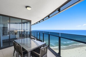 Ein Balkon mit Glastisch und Korbstühlen mit Blick auf einen Strand und das Meer.