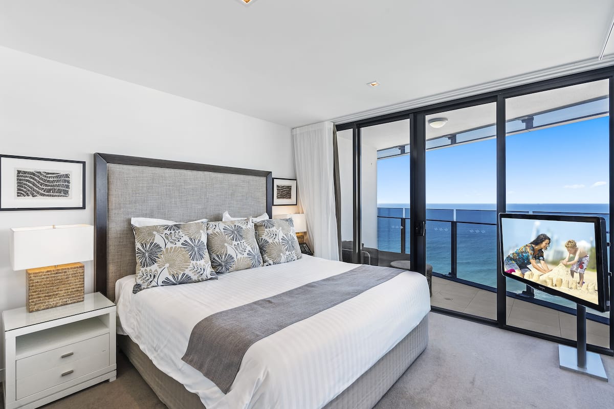 A modern bedroom with a large bed, a nightstand, a flat-screen TV, and a view of the ocean.