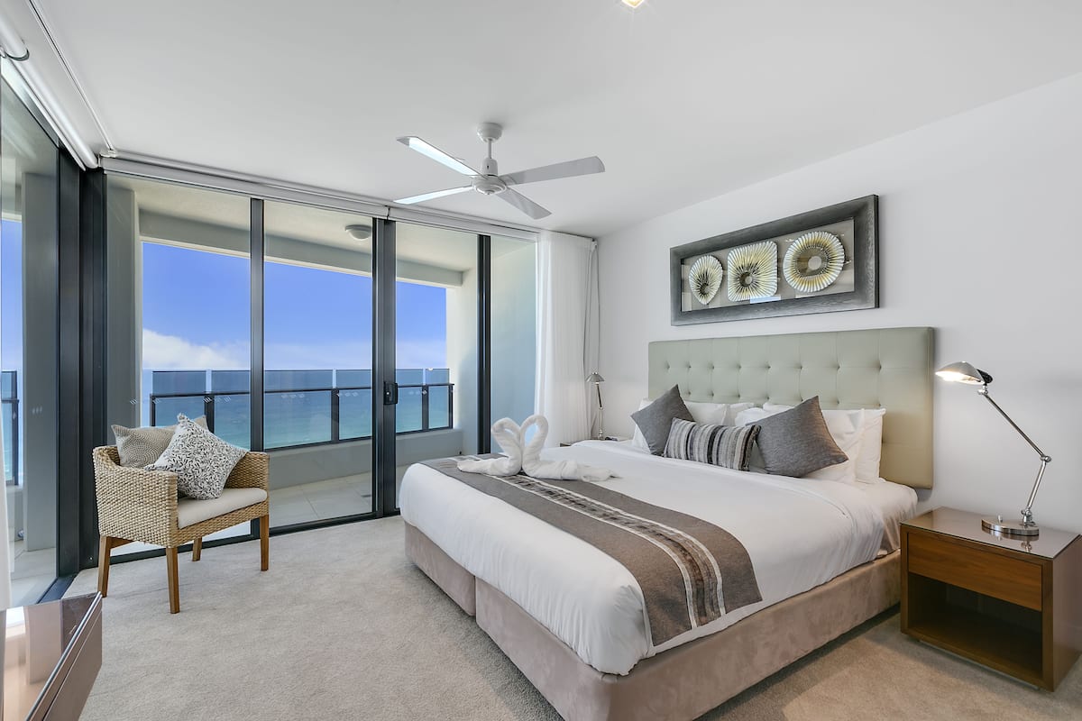 A modern bedroom with a large bed, a ceiling fan, a view of the ocean, and a balcony.