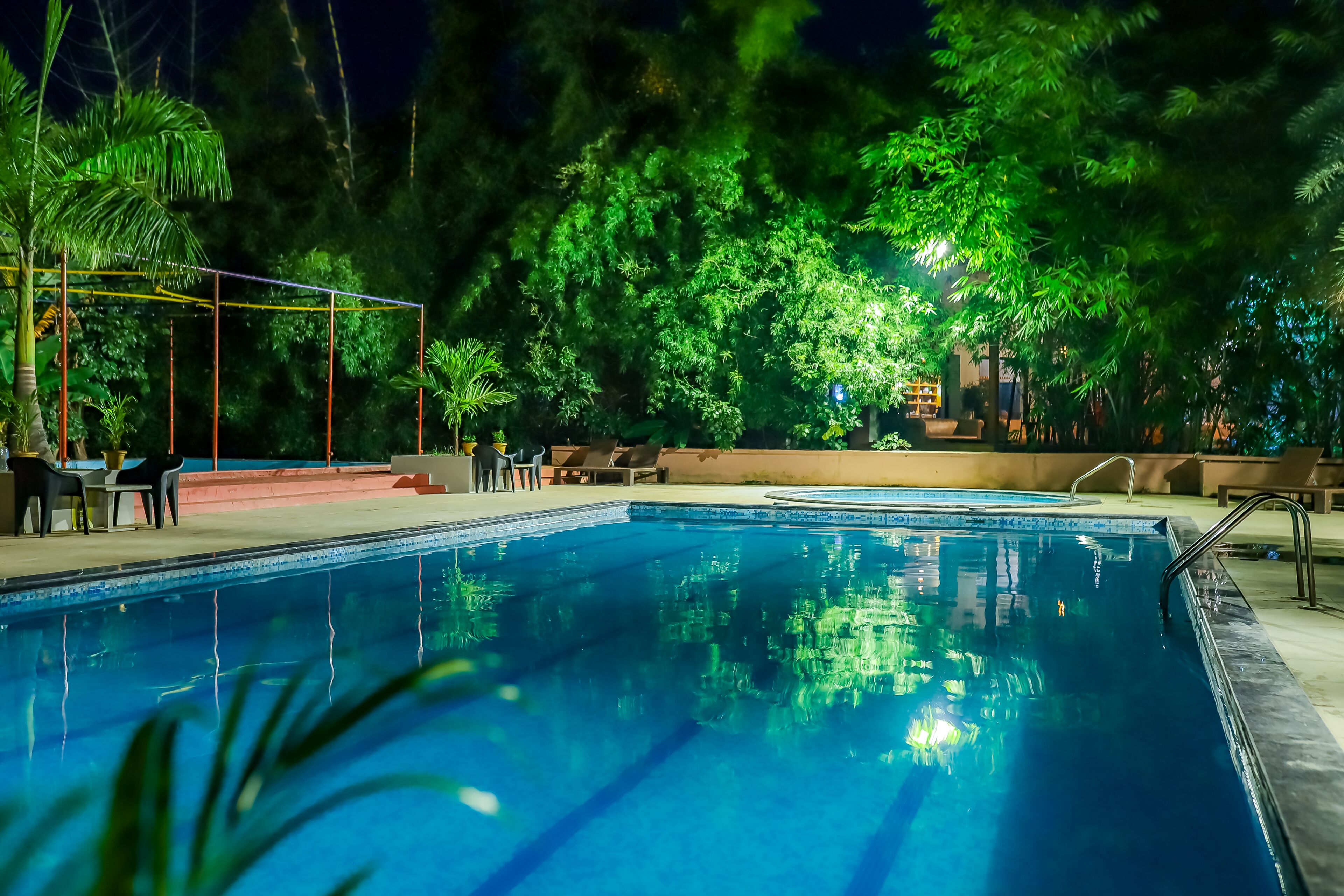 En swimmingpool omgiven av frodig grönska och träd, med ett väl upplyst däck och bekväma solstolar.