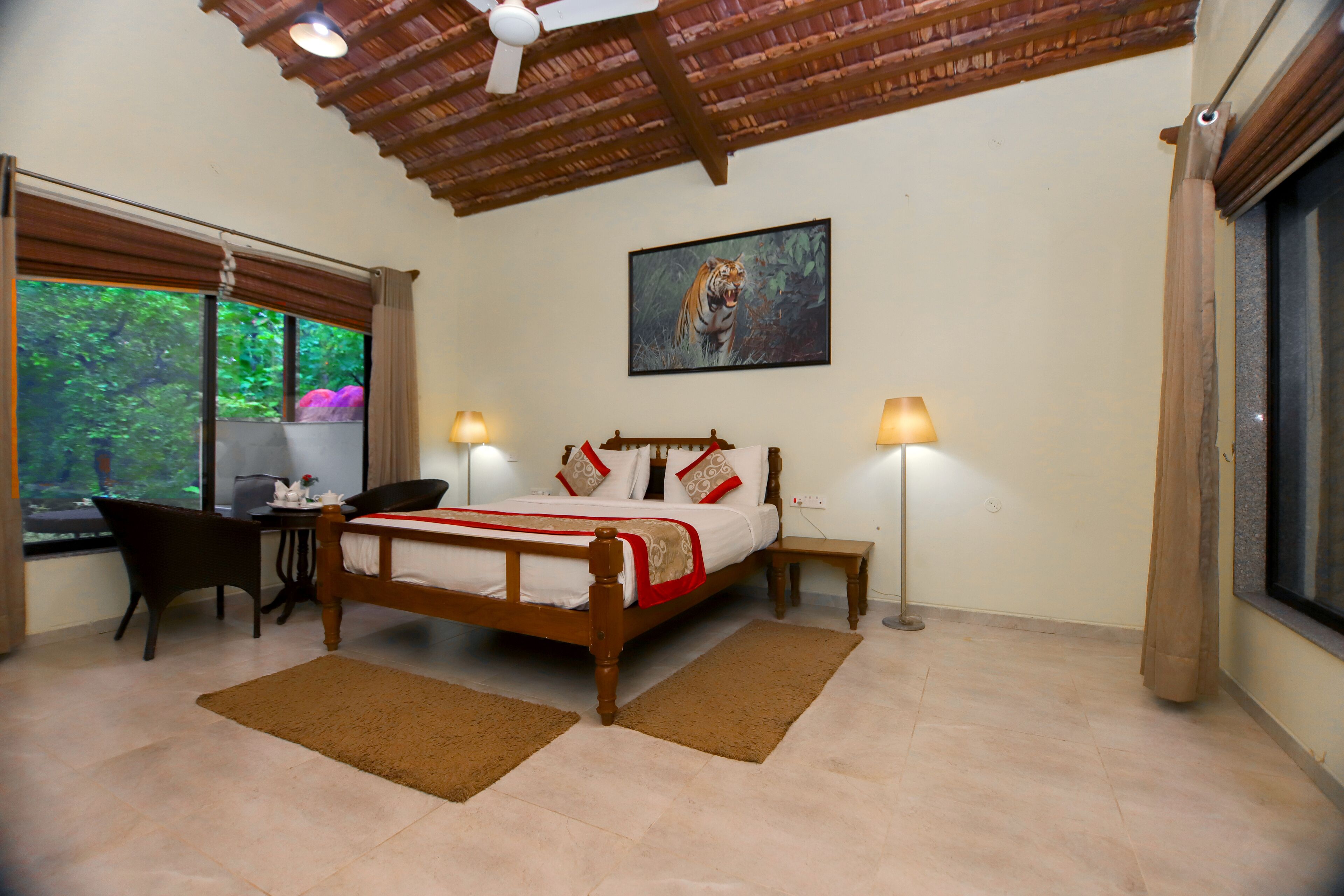 A bedroom with a wooden bed, two bedside tables with lamps, a desk with a chair, and a large window with a view of greenery.