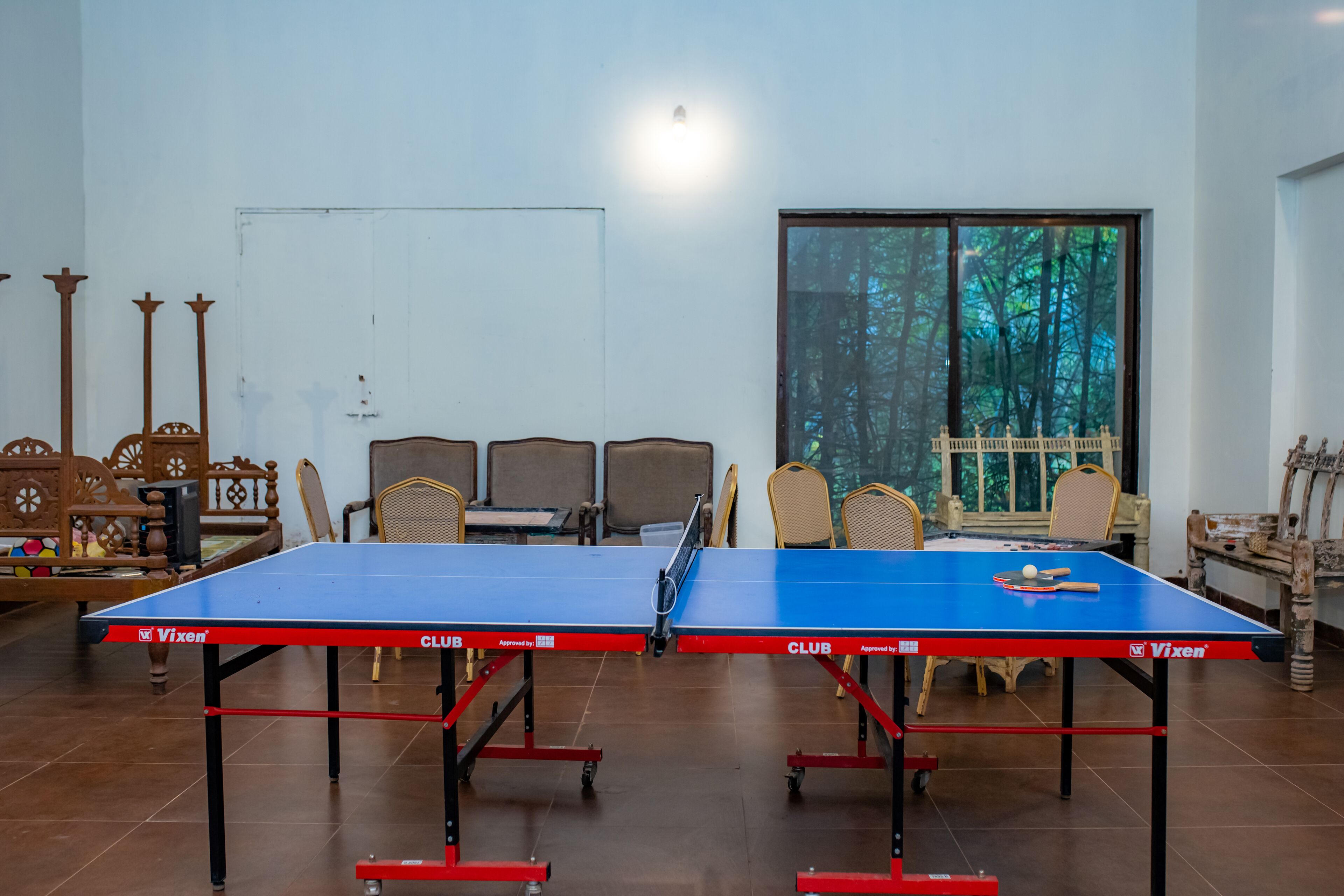 Una mesa de ping pong en una habitación con sillas, una pizarra blanca y un ventanal con vista a árboles.