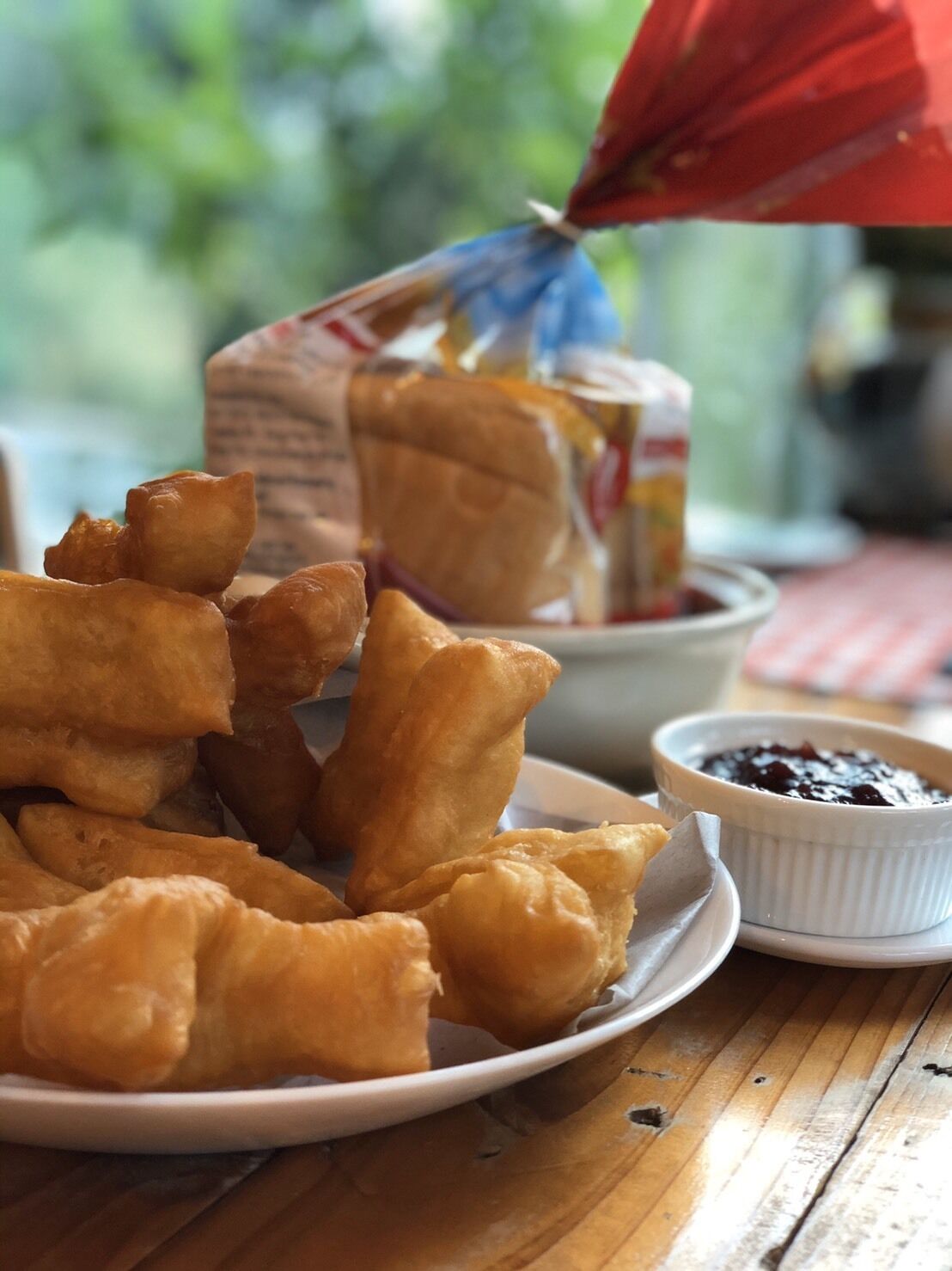 A plate of fried dough snacks with a small bowl of dark sauce, a loaf of bread, and a carton of milk on a wooden table.