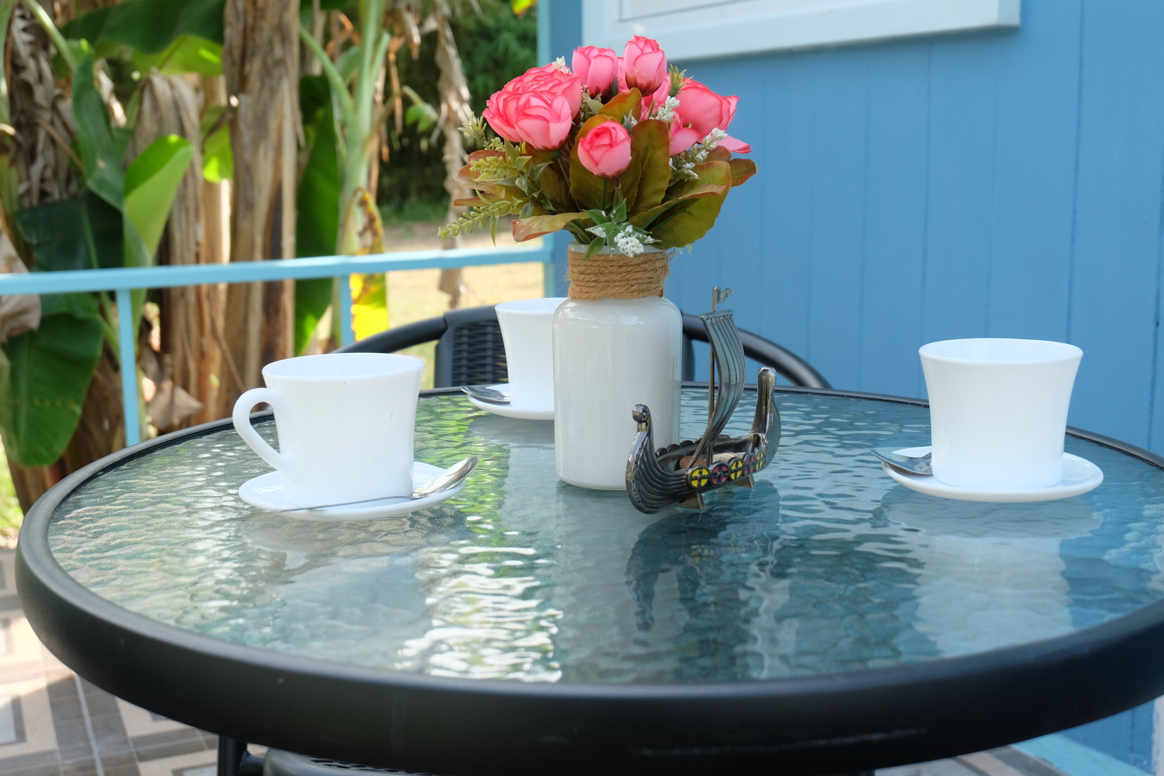 Una mesa de vidrio redonda con tazas y platillos blancos, un florero con flores rosadas y una figura decorativa de un pájaro.