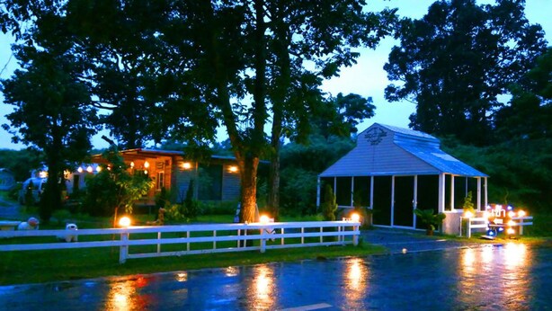 A covered pavilion with a roof, surrounded by a white fence, and a building with lights in the background. - Saengcha Farm Resort (Prachantakham)
