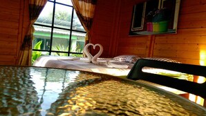 A wooden cabin room with a bed, a heart-shaped towel arrangement, a window with curtains, and a small shelf with potted plants. - Saengcha Farm Resort (Prachantakham)
