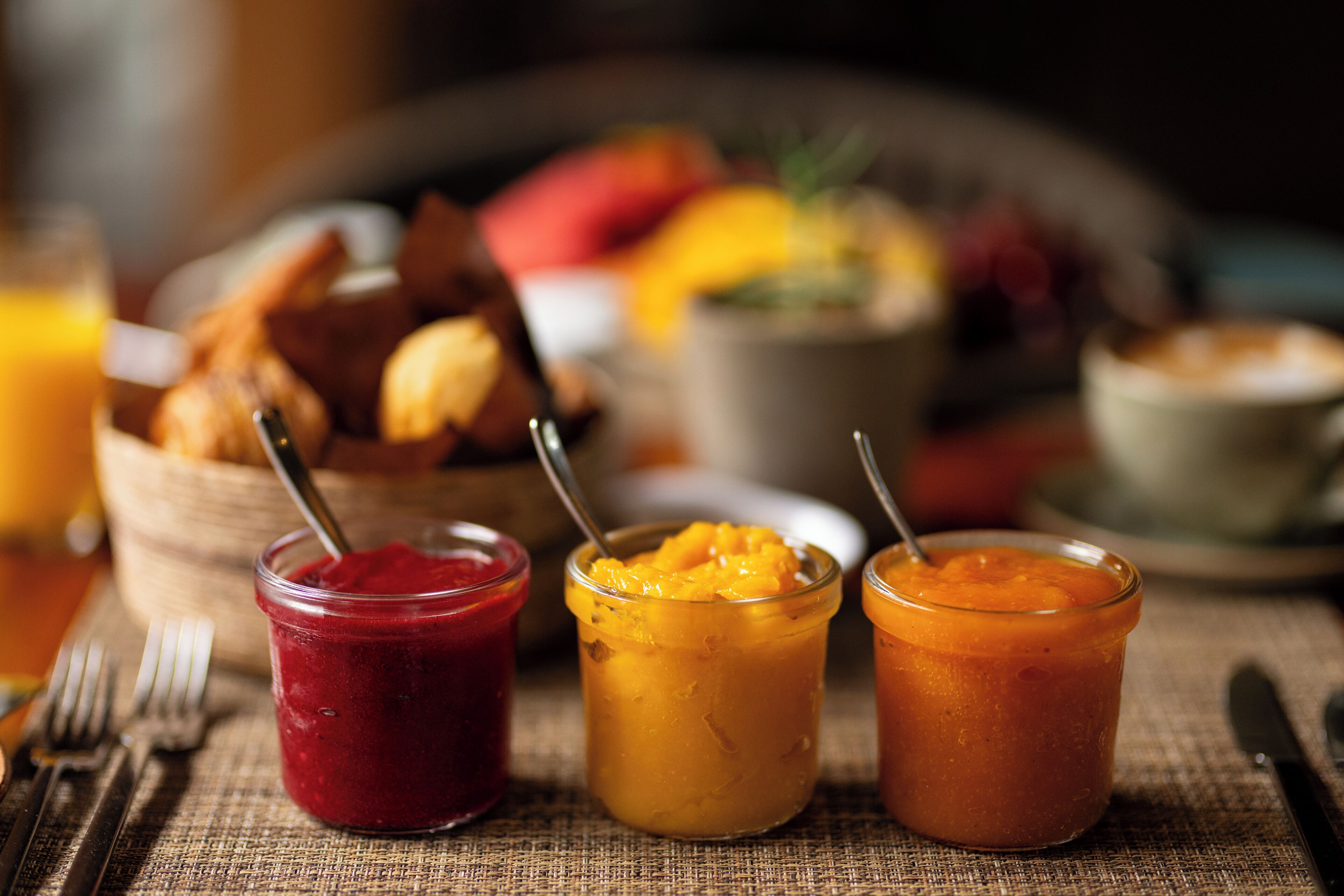 Trois pots de confitures de fruits sont posés sur une table, accompagnés d’un panier de pain et, en arrière-plan, on aperçoit des fruits et une tasse, le tout étant légèrement flou.