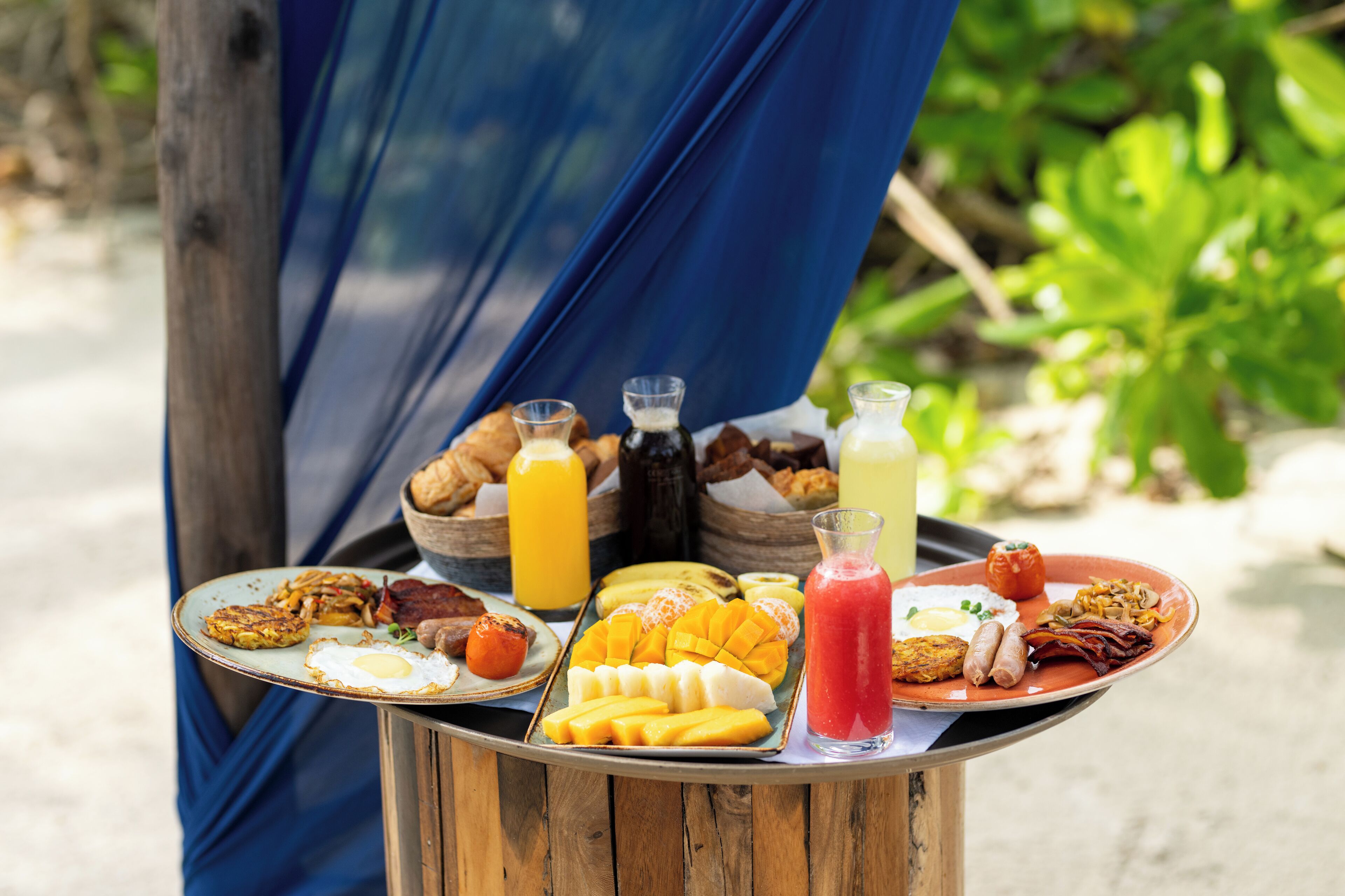 A breakfast spread on a wooden table outdoors with a blue cloth backdrop.