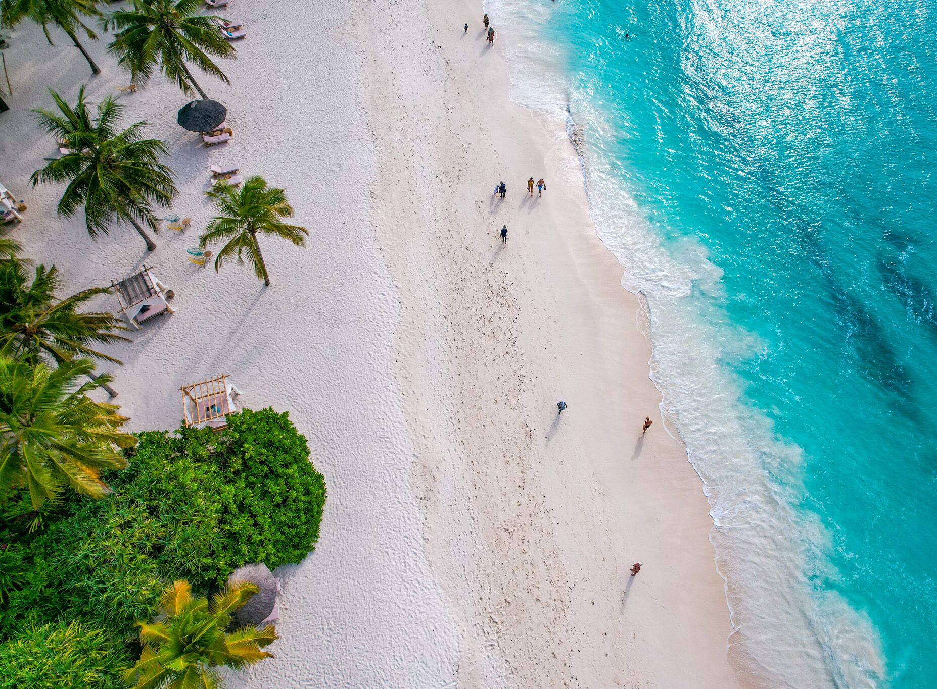 An aerial view of a beach with palm trees, beach chairs, and people walking along the shore.
