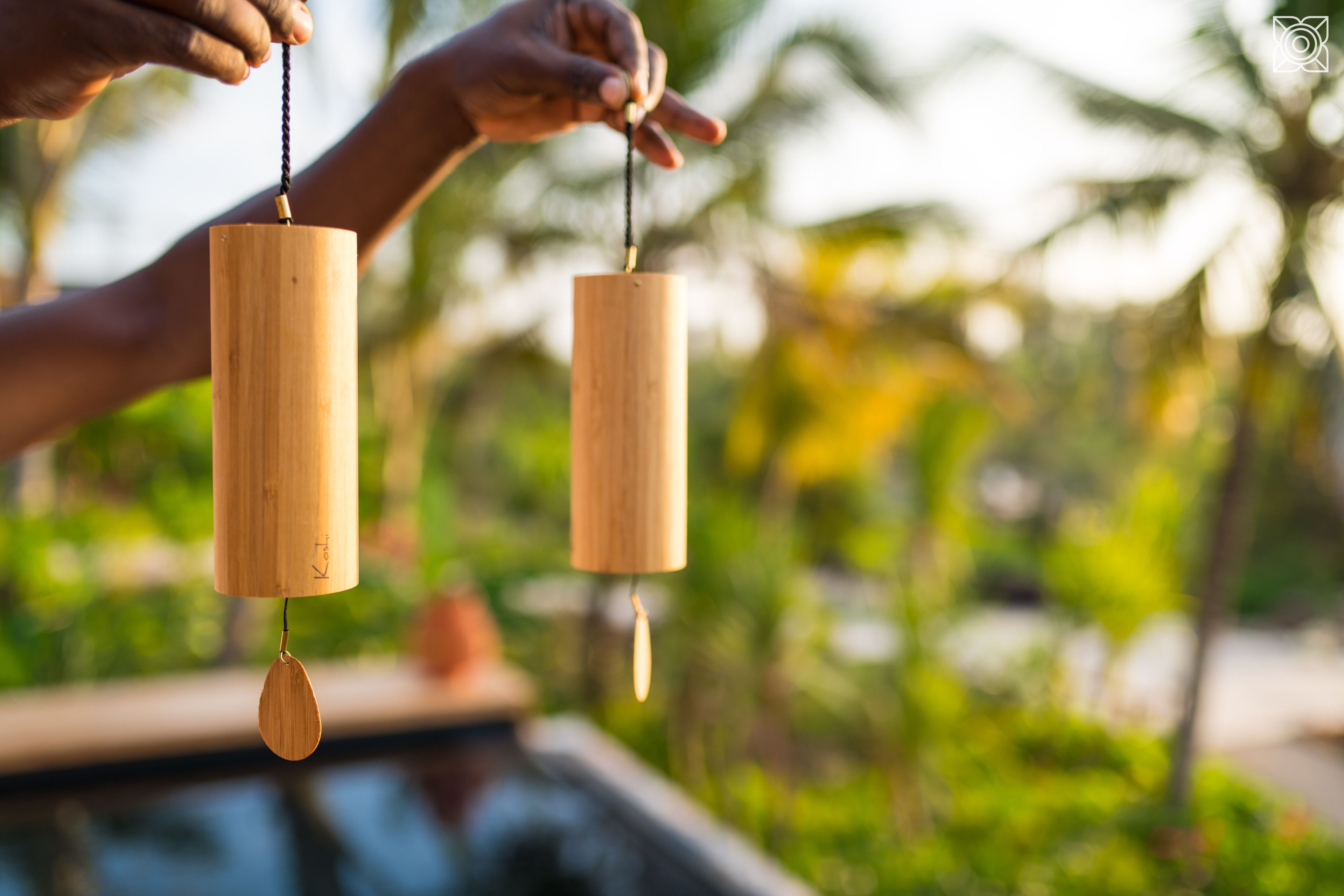Two wooden wind chimes hanging outdoors with a blurred background of greenery and a body of water.