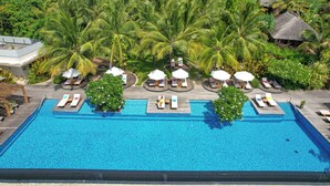 A large rectangular swimming pool with lounge chairs and umbrellas, surrounded by palm trees and thatched-roof huts.