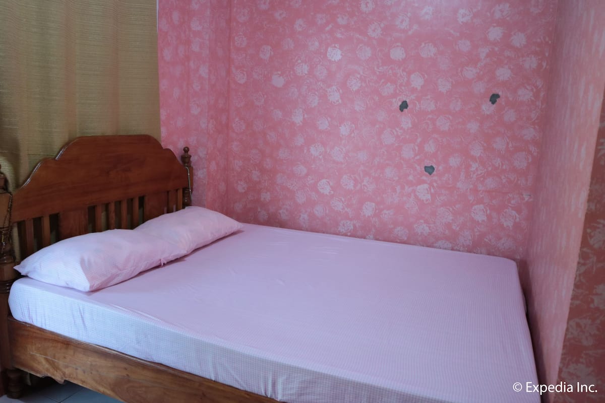 Un lit simple avec une tête de lit en bois, dans une pièce dont le papier peint est orné de motifs floraux roses.