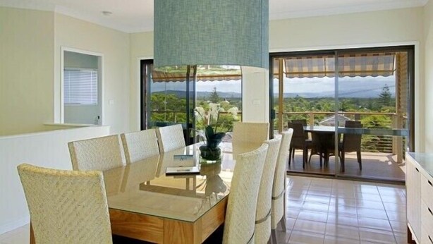 A dining area with a table and chairs, a hanging light fixture, and a view of greenery through large windows. - Away on Massinger Street (Byron Bay)