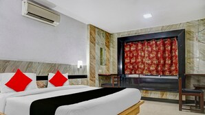 A hotel room with a bed, a window with a red curtain, a small table, and an air conditioning unit. - Hotel Arch Manor Deluxe (Bhopal)