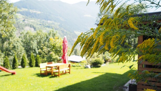 A garden with a wooden bench and a red umbrella, surrounded by greenery and mountains in the background. - Bei Herrmanns (Sautens)