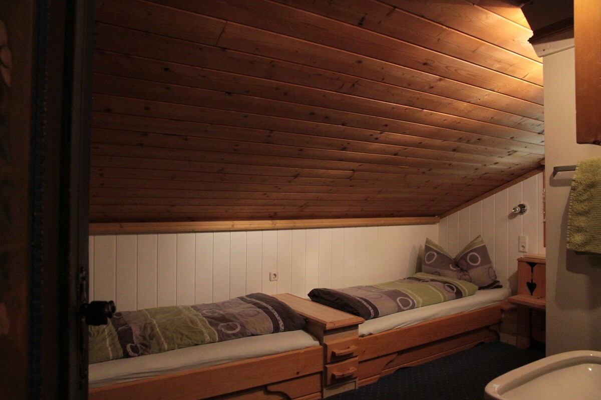 A small, cozy room with a wooden ceiling and walls, a single bed with a patterned bedspread, a small wooden desk, and a white toilet.