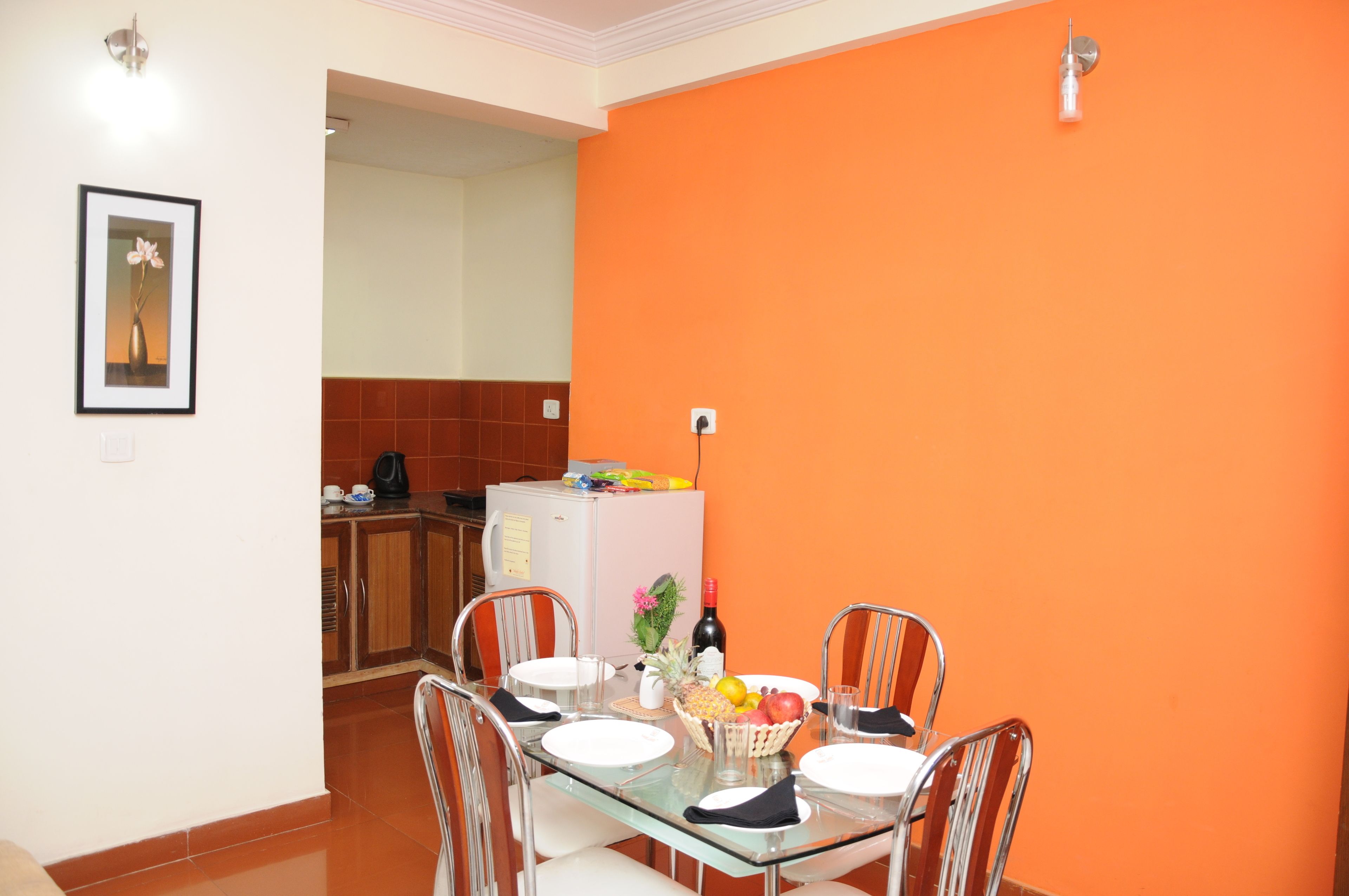 Un comedor con una mesa de vidrio, sillas metálicas y un frutero. Se observan un refrigerador y armarios de cocina al fondo.