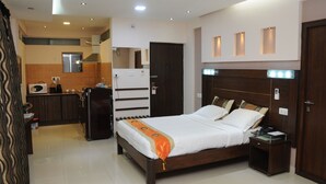 A hotel room with a bed, bedside table, and a small desk with a lamp. There is a kitchen area with a refrigerator and microwave. - Maple Suites (Bengaluru)