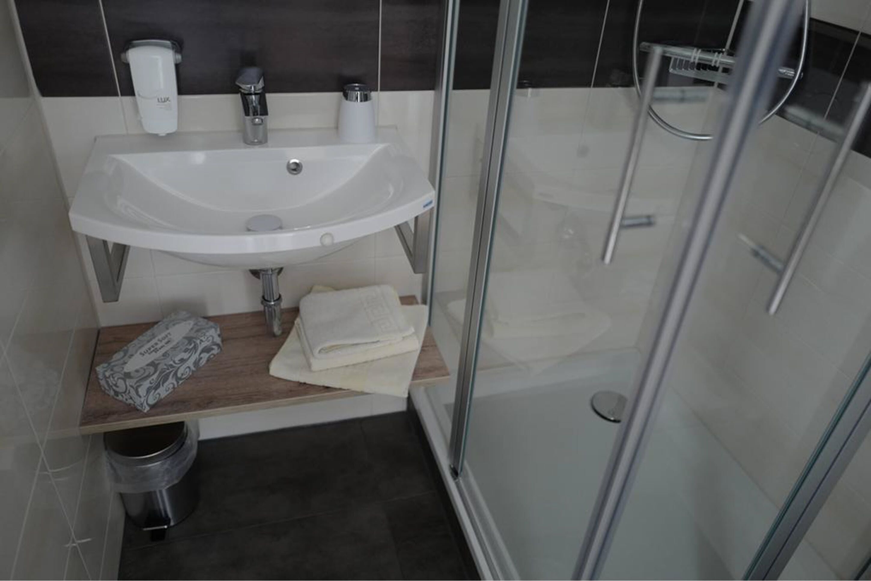 Un baño moderno con ducha de vidrio, un lavabo blanco y un pequeño estante de madera con toallas.