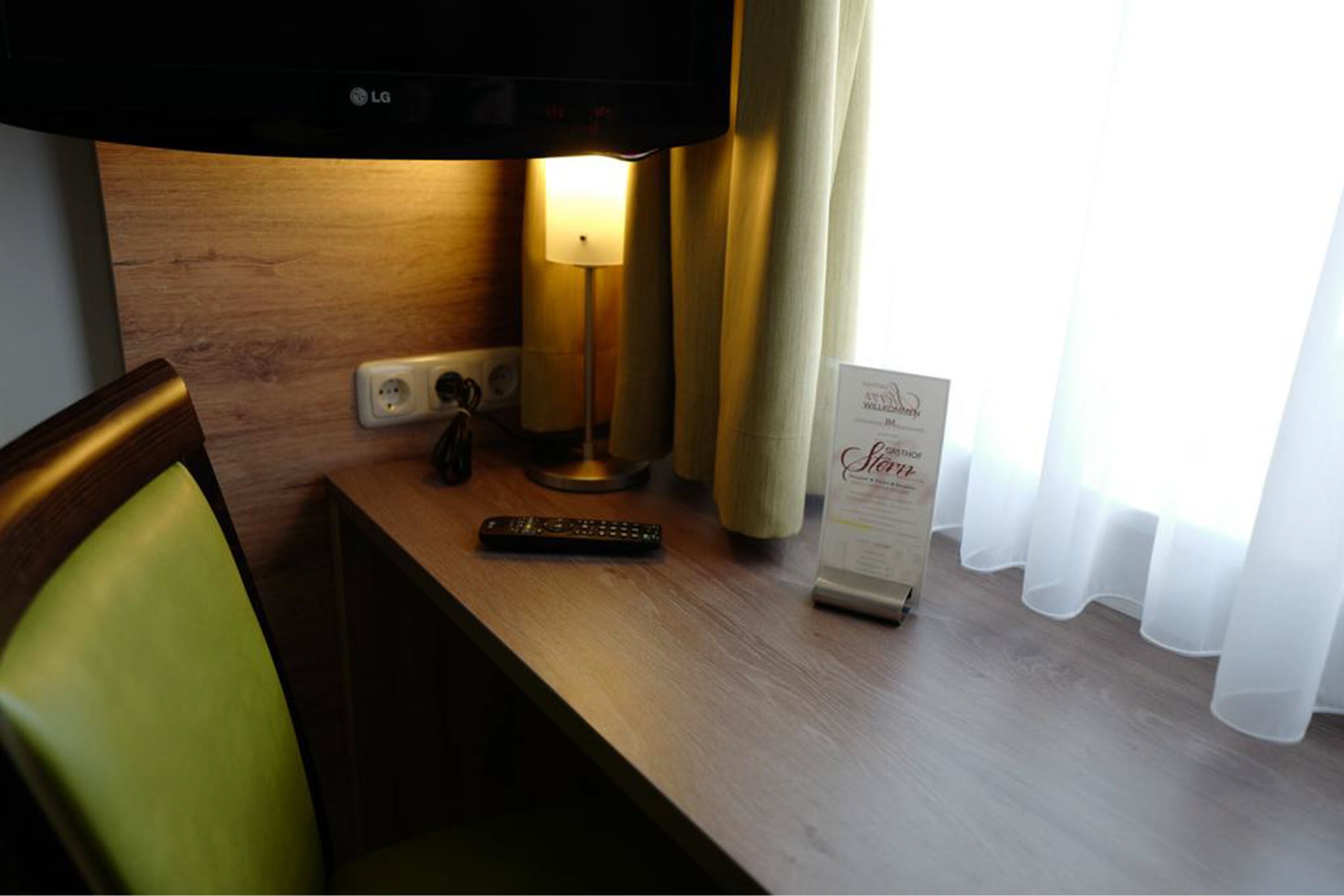 A hotel room with a wooden desk, a lamp, a remote control, a sign, and a chair.