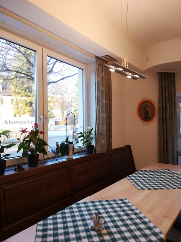 A dining area with a large window, wooden table, checkered tablecloths, and potted plants.