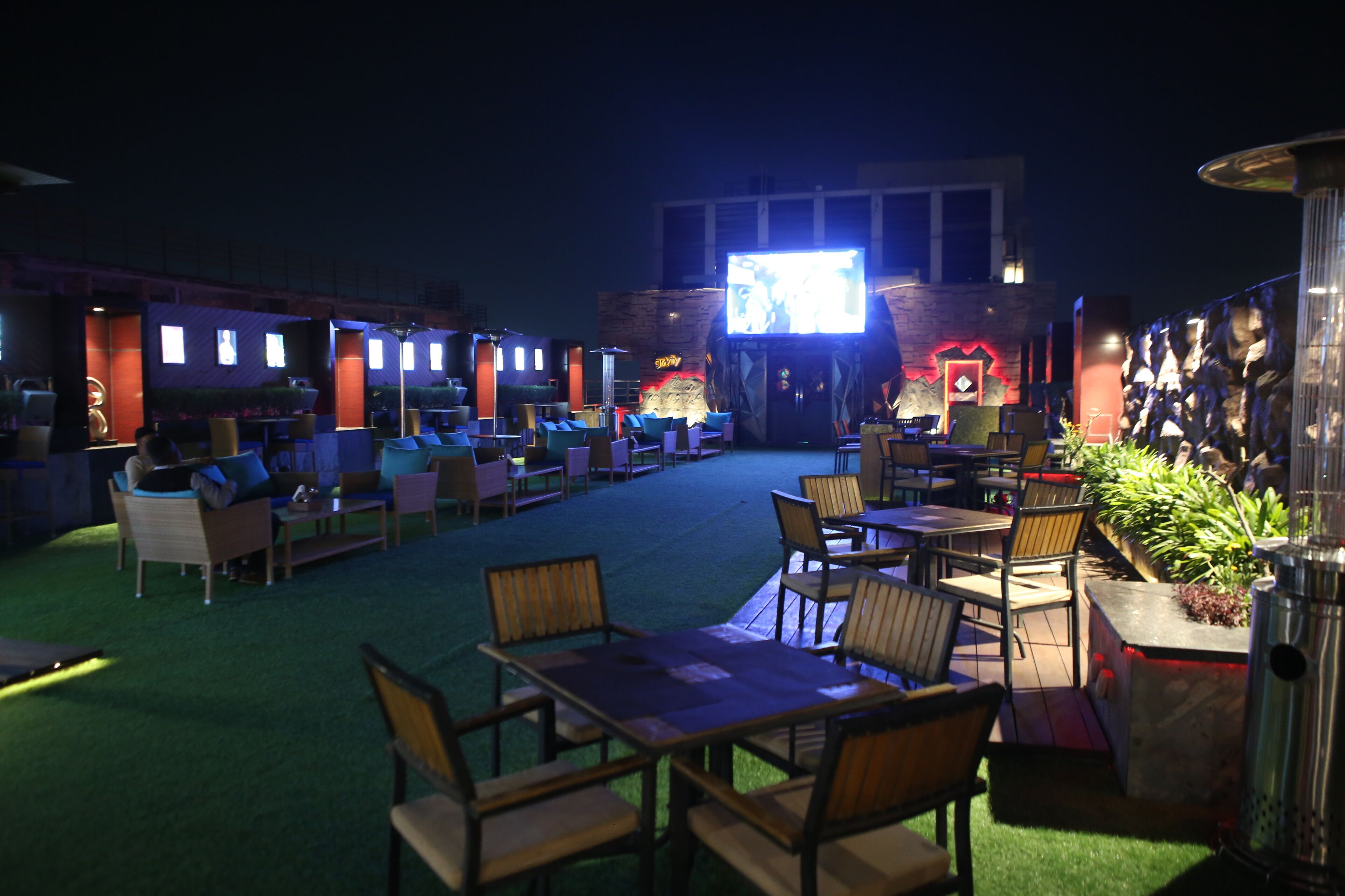 An outdoor seating area with tables and chairs, a TV screen, and a stone wall with a neon sign.