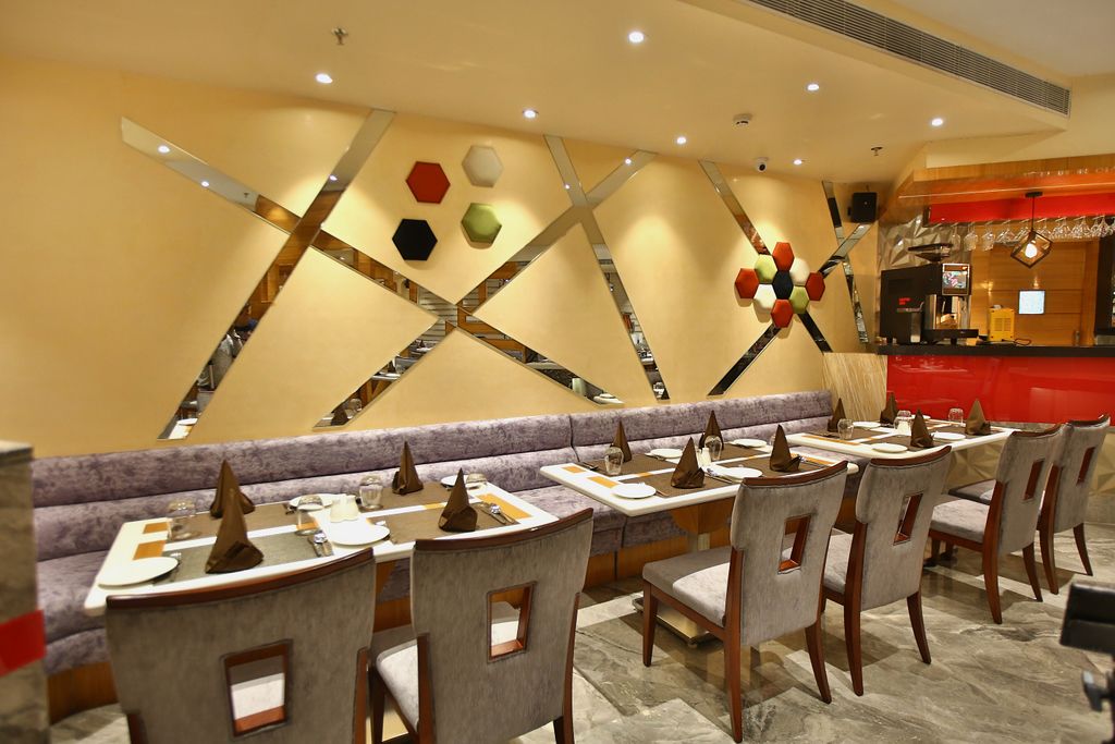 A restaurant with a modern interior, featuring a long counter with multiple tables and chairs, a decorative wall with geometric patterns, and a red accent wall with a coffee machine.