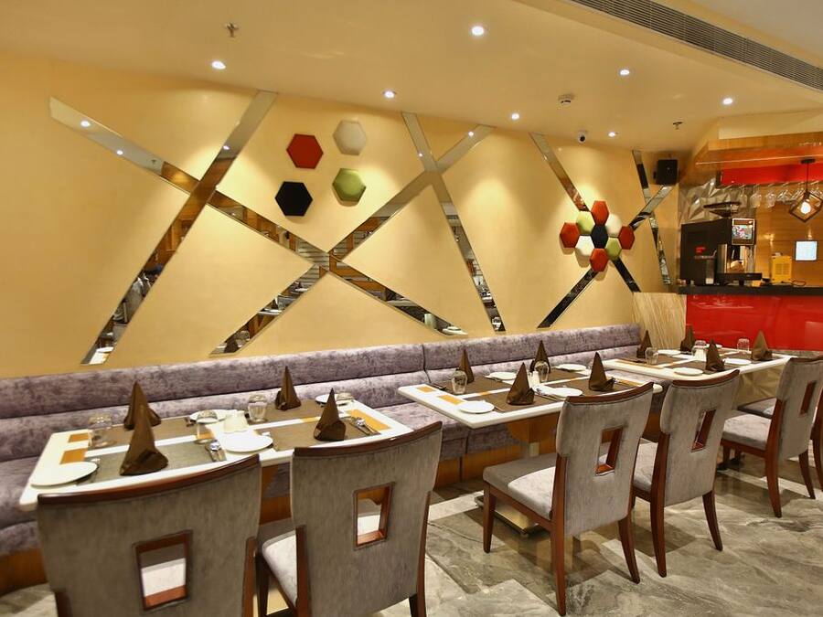 A restaurant with a modern interior, featuring a long counter with multiple tables and chairs, a decorative wall with geometric patterns, and a red accent wall with a coffee machine.