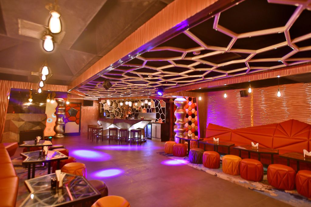 A modern bar area with hexagonal ceiling design, seating with orange cushions, and a bar counter with stools.