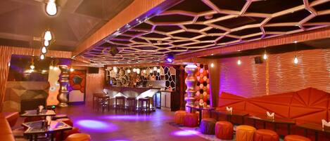 A modern bar area with hexagonal ceiling design, seating with orange cushions, and a bar counter with stools.