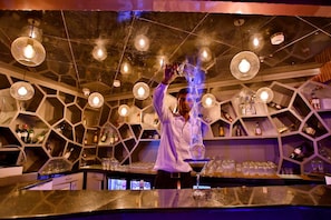 Zona de bar con estantes hexagonales llenos de botellas, un barman sirviendo una bebida y lámparas colgantes.