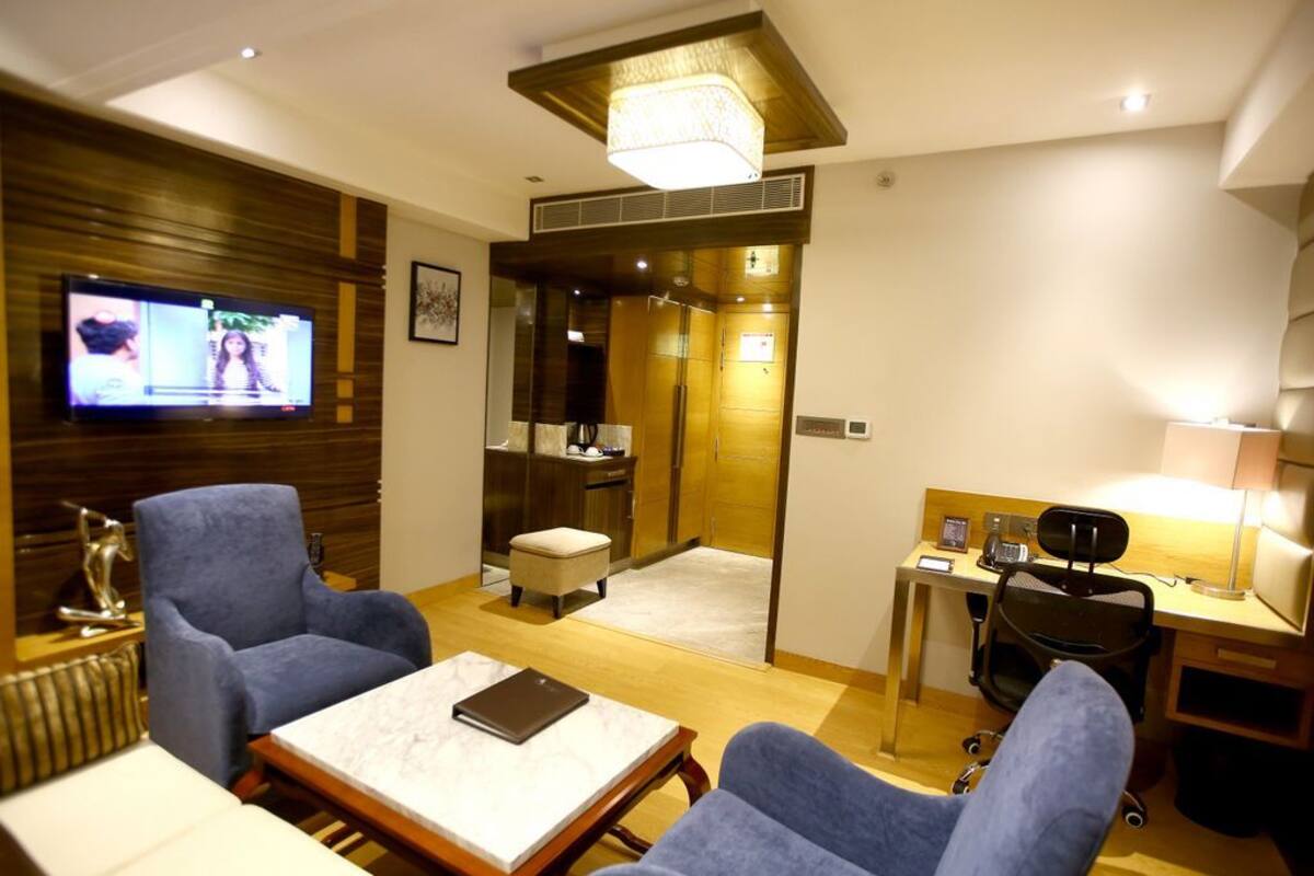 A hotel room with a television, two armchairs, a small table, a desk with a chair, and a bathroom visible through a glass door.