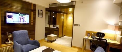 A hotel room with a television, two armchairs, a small table, a desk with a chair, and a bathroom visible through a glass door.