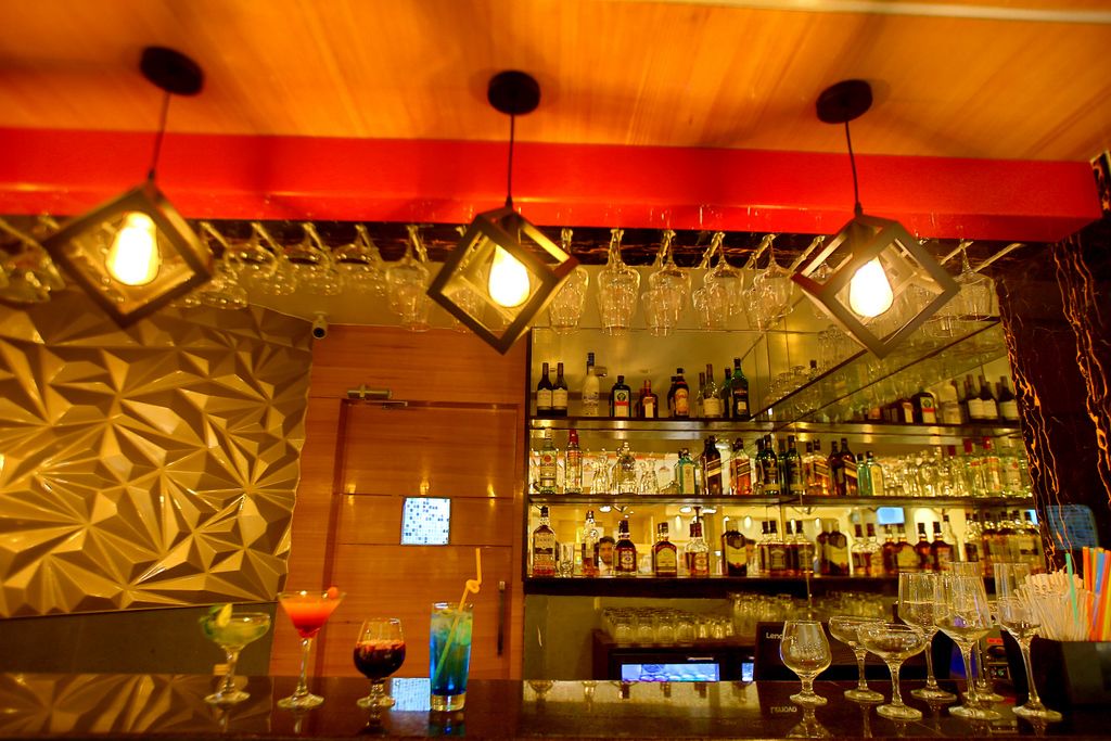 Eine Bar mit hängenden Lampen, einer Auswahl an Flaschen und Gläsern auf Regalen sowie einer Wand mit Muster.