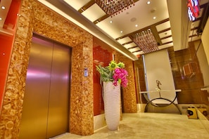Un ascensor moderno con diseño de pared de mármol, un jarrón grande con flores y una lámpara contemporánea.