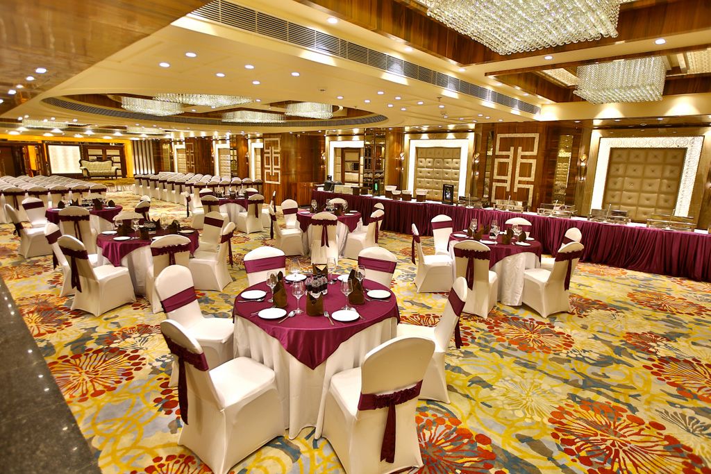 Une salle de banquet avec des tables rondes, des chaises blanches ornées de rubans violets et un sol recouvert de moquette.