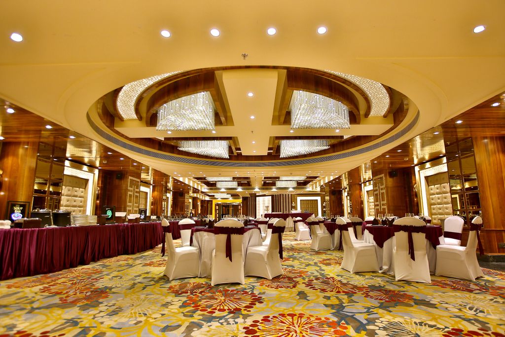 Une grande salle de banquet, bien éclairée, avec un plafond à motifs circulaires, des rangées de chaises blanches ornées de rubans rouges et une moquette aux couleurs vives.