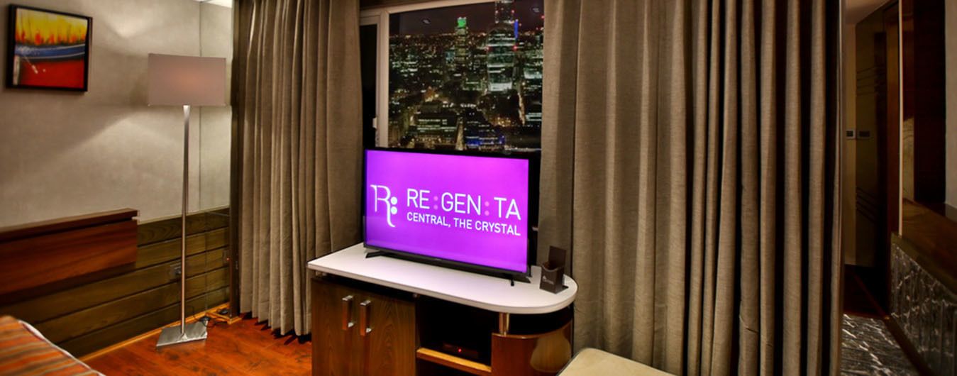 A hotel room with a television displaying "RE:GEN:TA CENTRAL, THE CRYSTAL" on the screen, a cityscape view through the window, and a wooden cabinet with drawers.