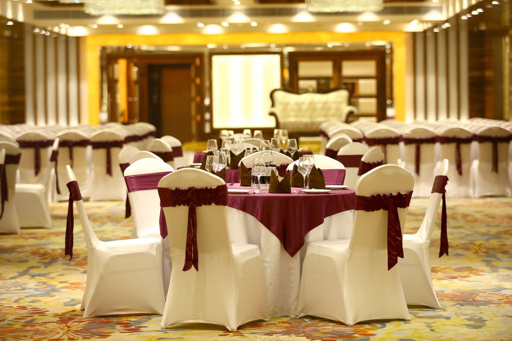 A banquet hall with round tables set for a formal event, white chairs with maroon sashes, and a carpeted floor.