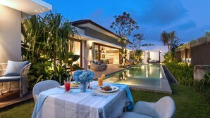A poolside dining area with a table set for two, surrounded by lush greenery and a modern house in the background. - Amarin Seminyak (Seminyak)