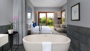 A modern hotel room with a large bathtub, a bed with white linens, a sofa, and a view of a garden through the sliding doors. - Amarin Seminyak (Seminyak)