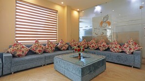 A modern living room with a grey sofa adorned with floral cushions, a glass coffee table with a vase of flowers, and a large window with blinds. - Jagdish Residency (Reasi)